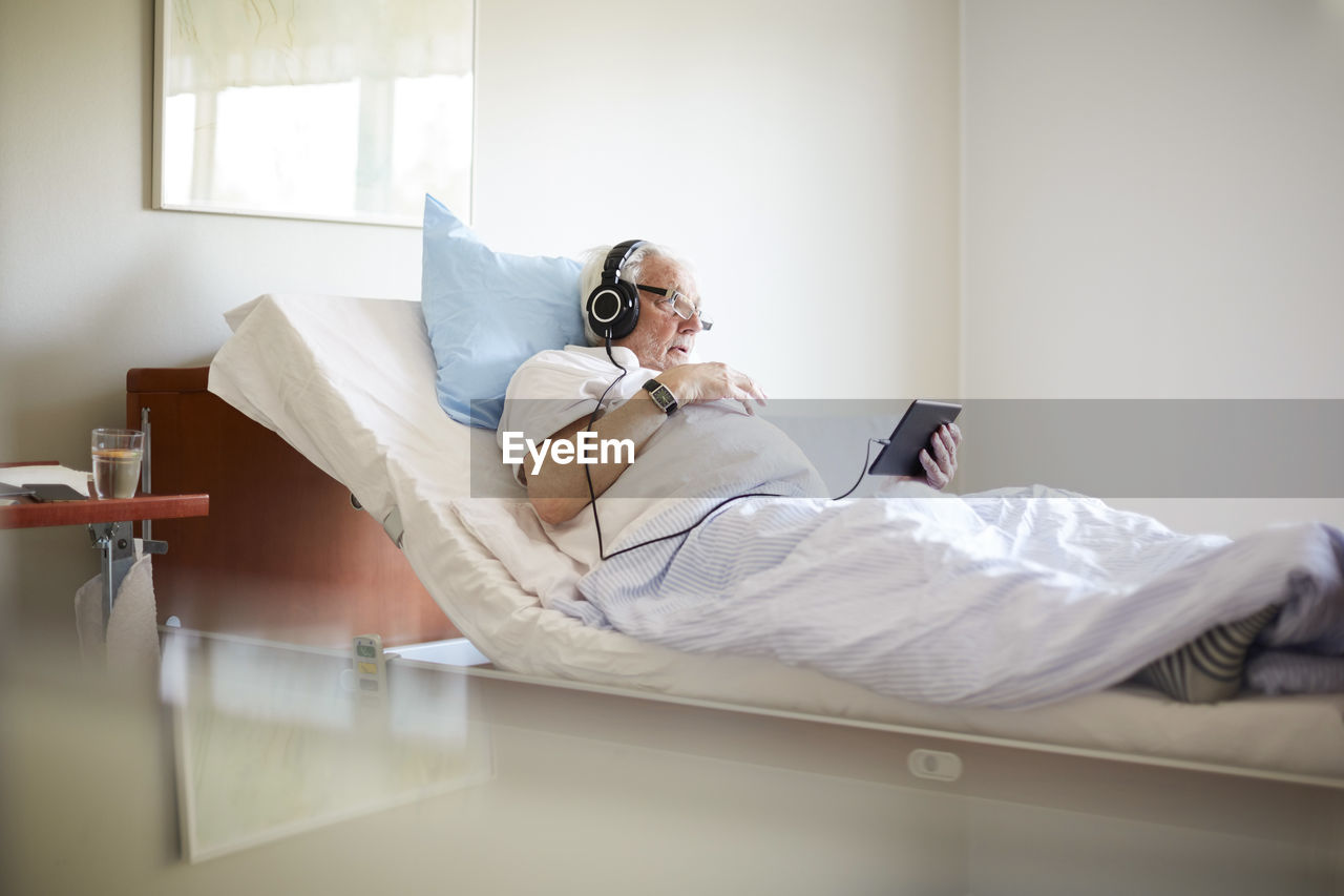 Senior man using digital tablet on bed in hospital ward