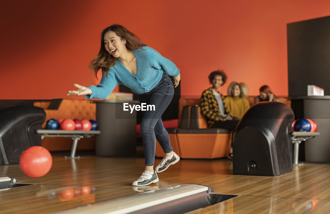 Young woman throwing ball with friends sitting in background at bowling alley