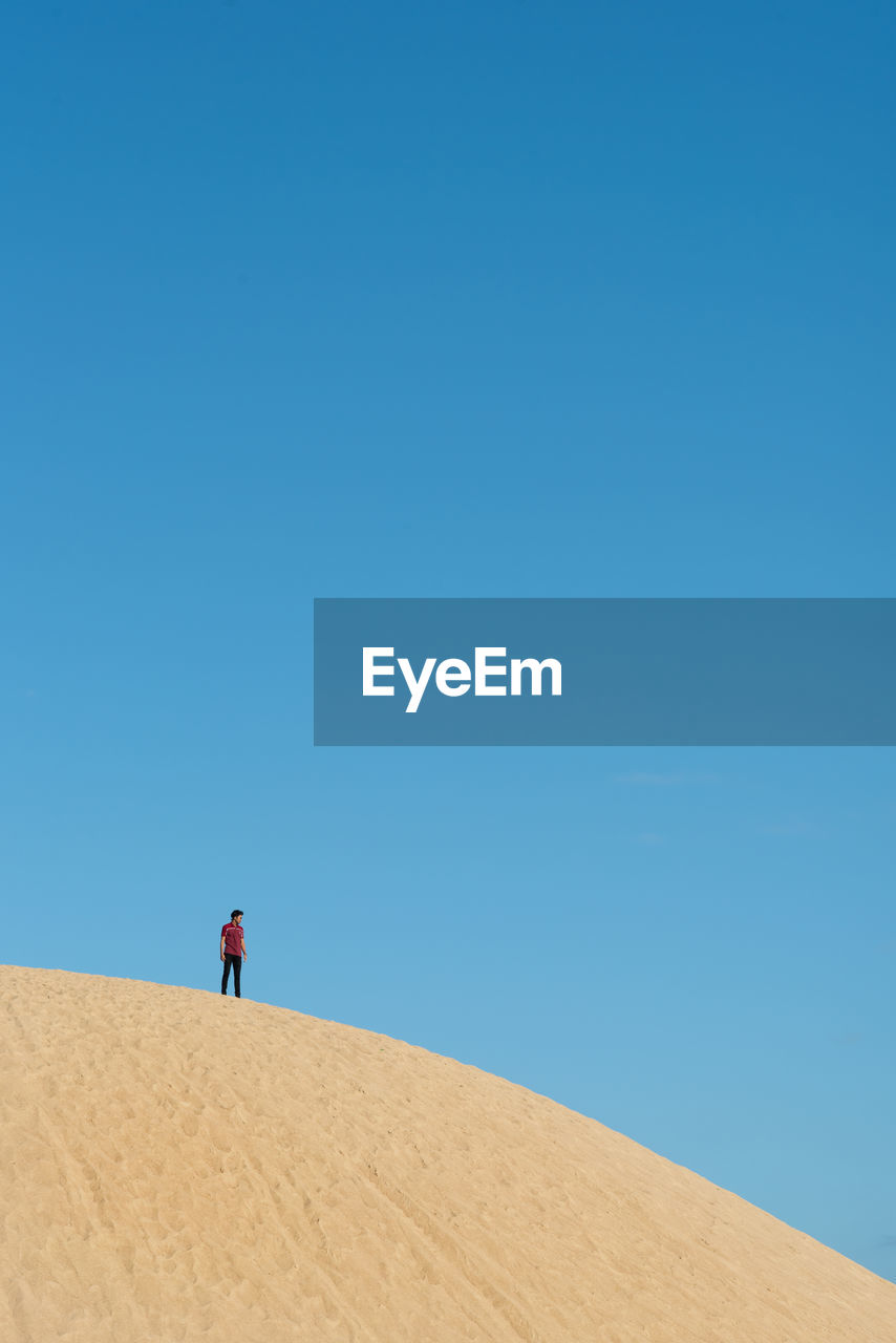 Low angle view of man standing on sand dune against blue sky