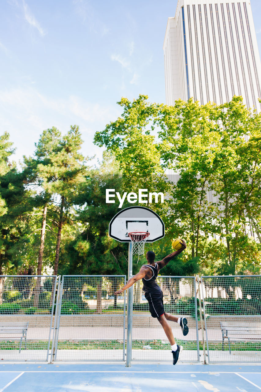 Back view of full body african american sportsman jumping and throwing ball in basketball hoop on sports ground in sunny day