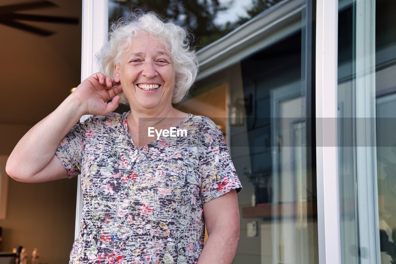 A happy elderly grey-haired woman smiles at the doorway of her house.