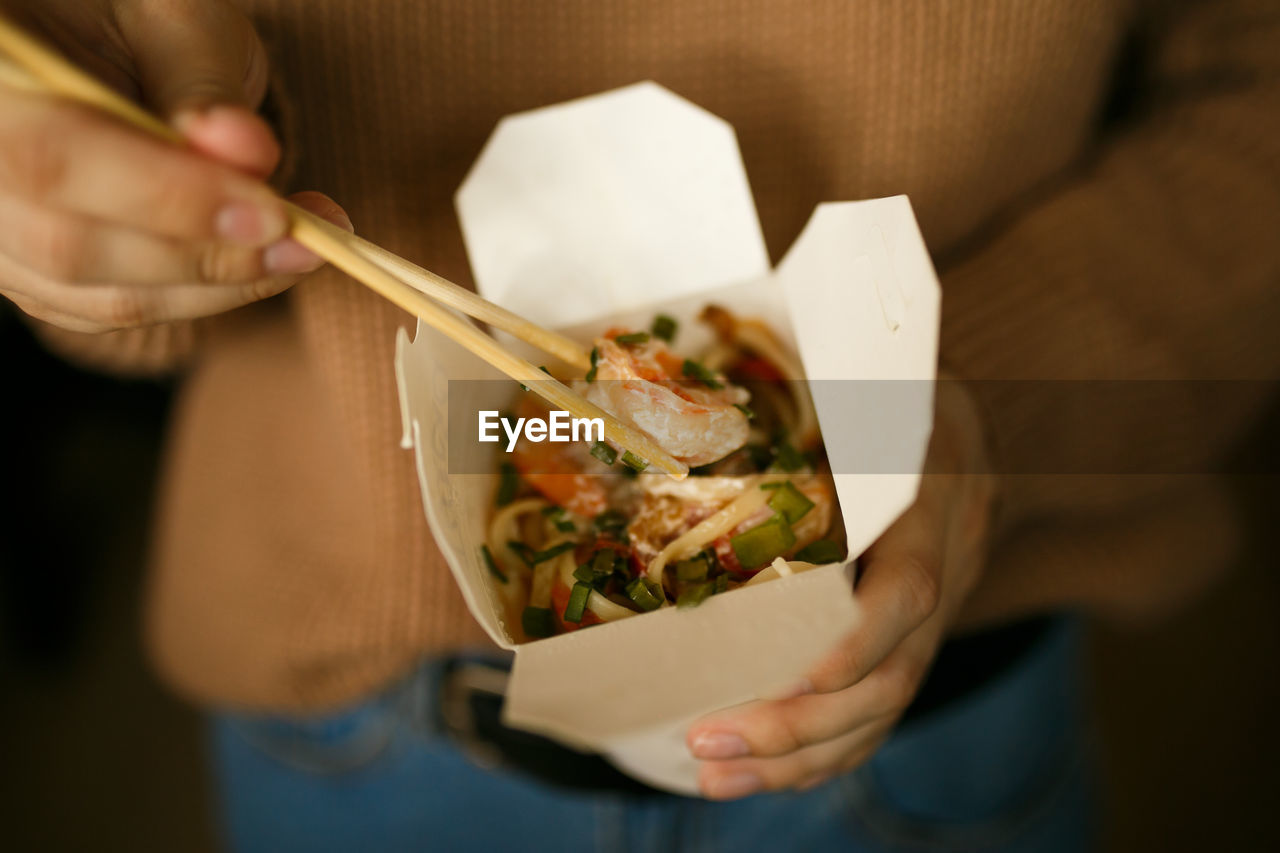 Girl in casual clothes eating noodles from a box sitting on the floor of the house, food delivery