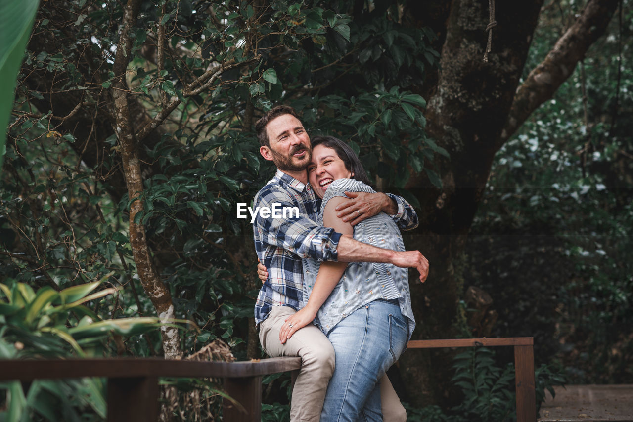 Cheerful couple in love embracing on wooden terrace of house in woods and laughing