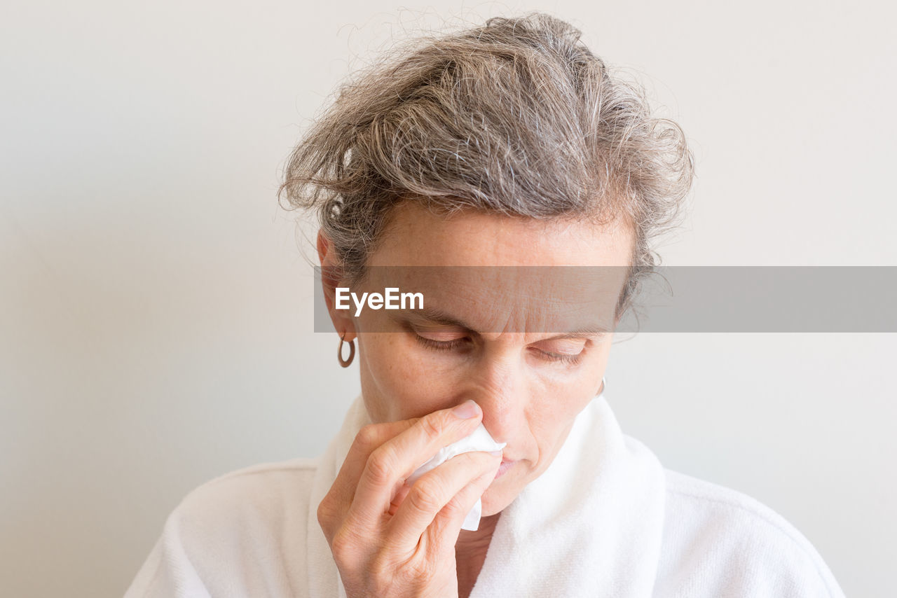 Close-up of woman cleaning nose with facial tissue against wall