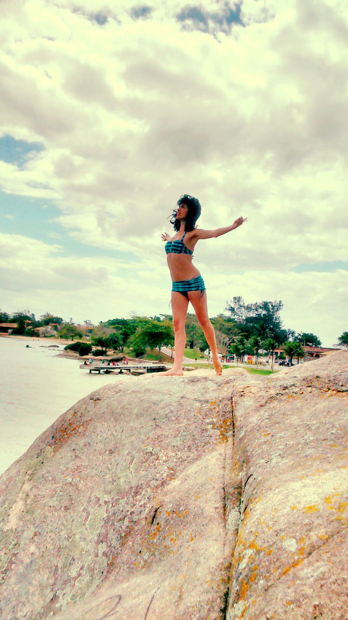 Full length of woman in bikini standing on rock at beach against cloudy sky