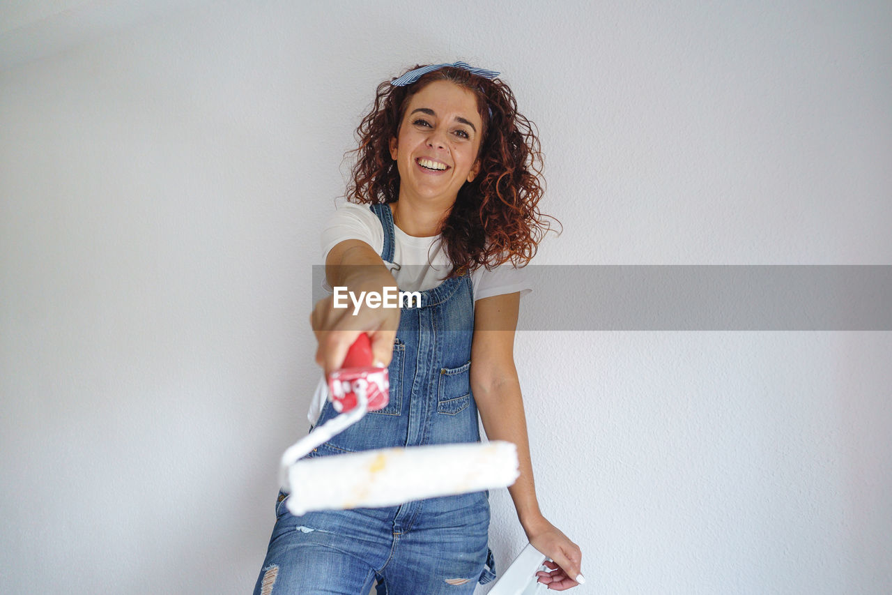 Portrait of smiling woman holding paint roller while standing against wall