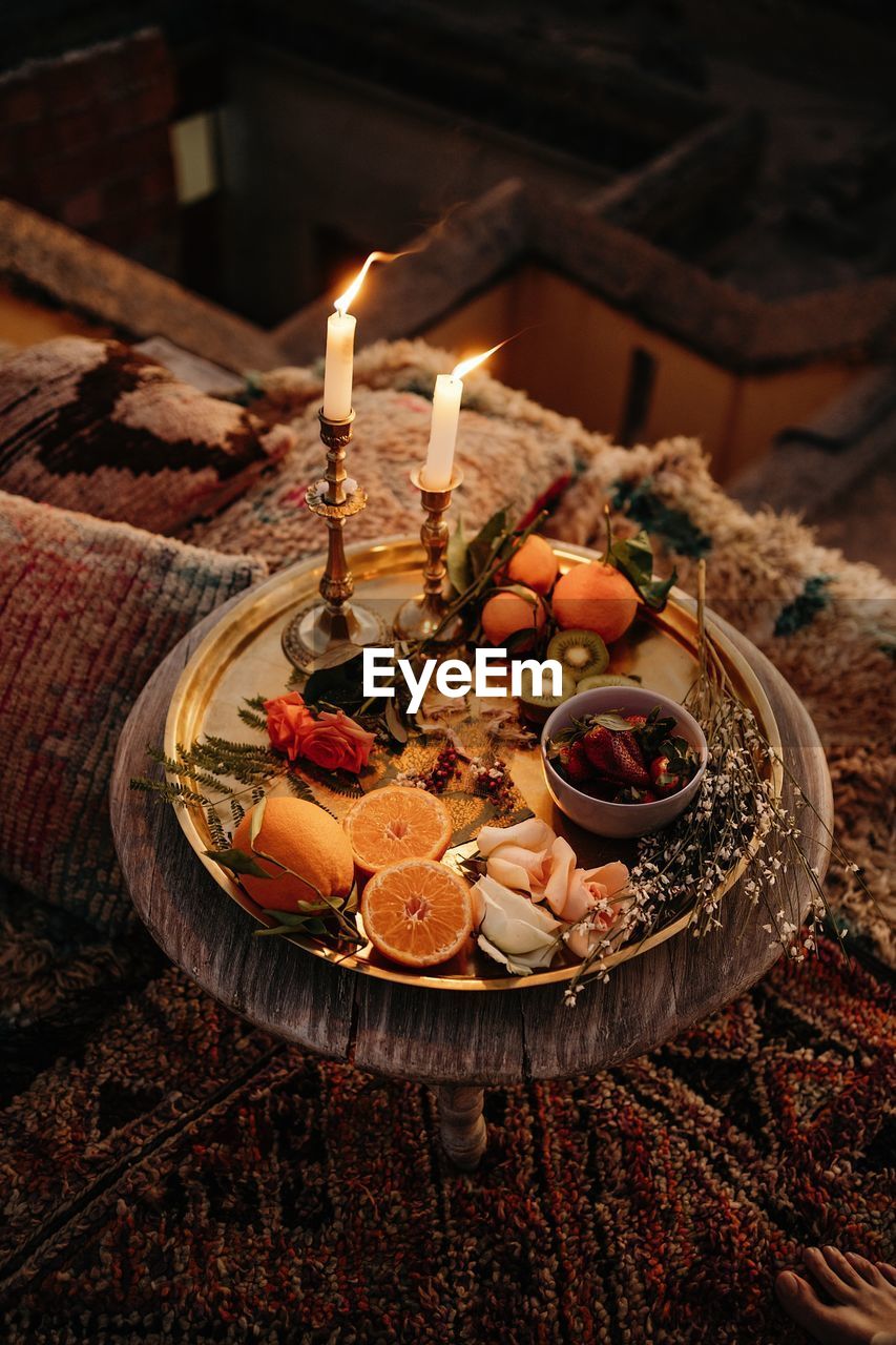 From above composition of assorted fresh fruits and plants served on golden tray with burning candles on round wooden table in moroccan style