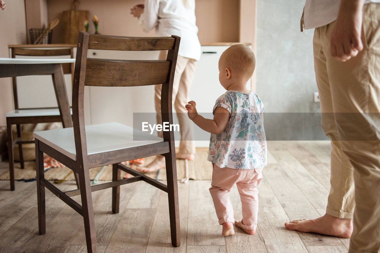 Little baby walks by wooden floor in kitchen between parents legs.