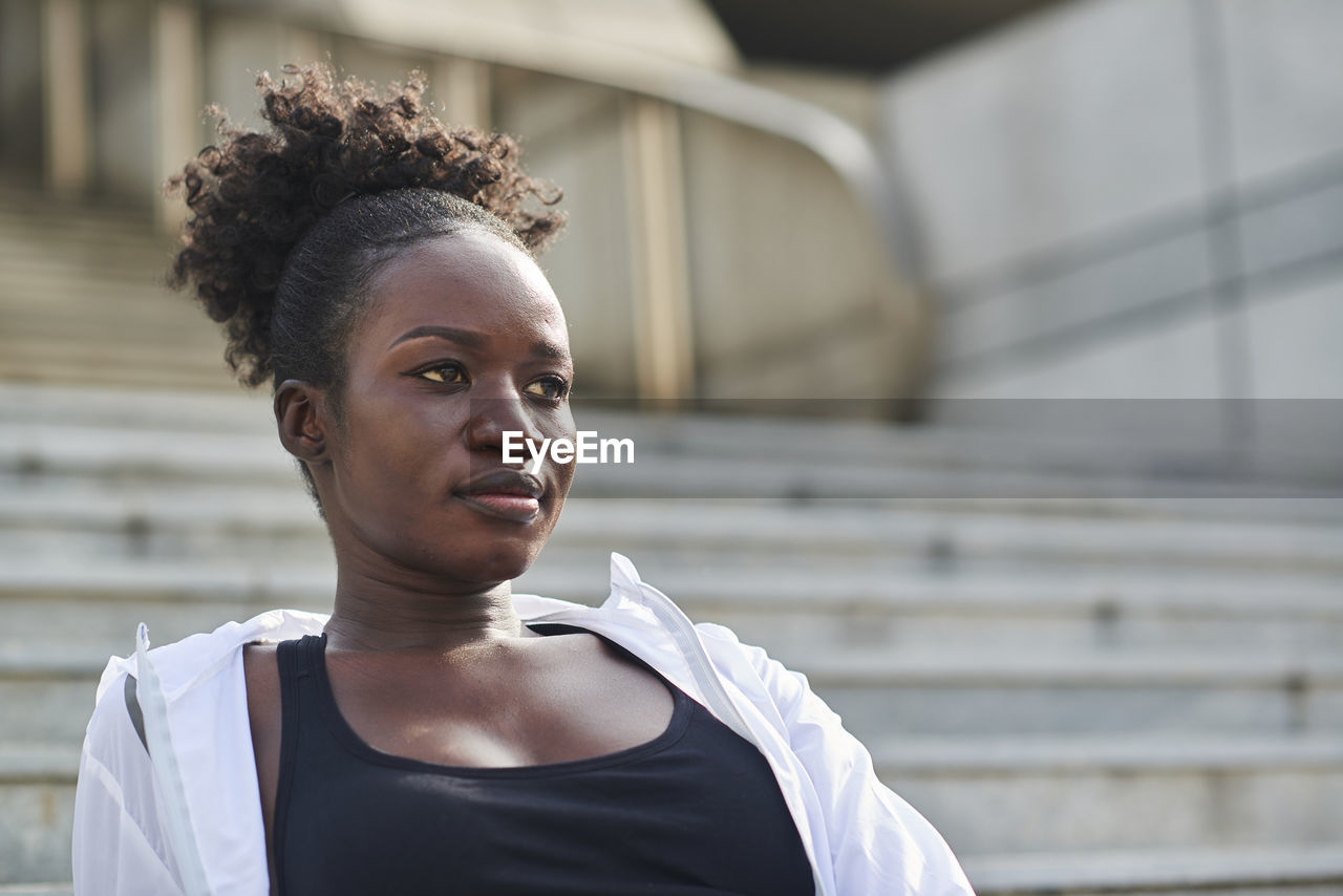 Confident african american female runner sitting on stone stairs on street while relaxing during workout and looking away