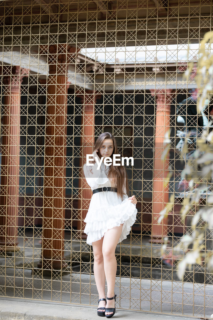 Full length portrait of young woman standing against metal grate