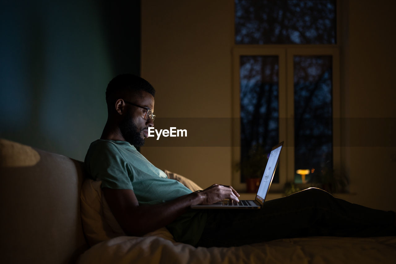 Concentrated african american programmer man lying on bed work on laptop at night.