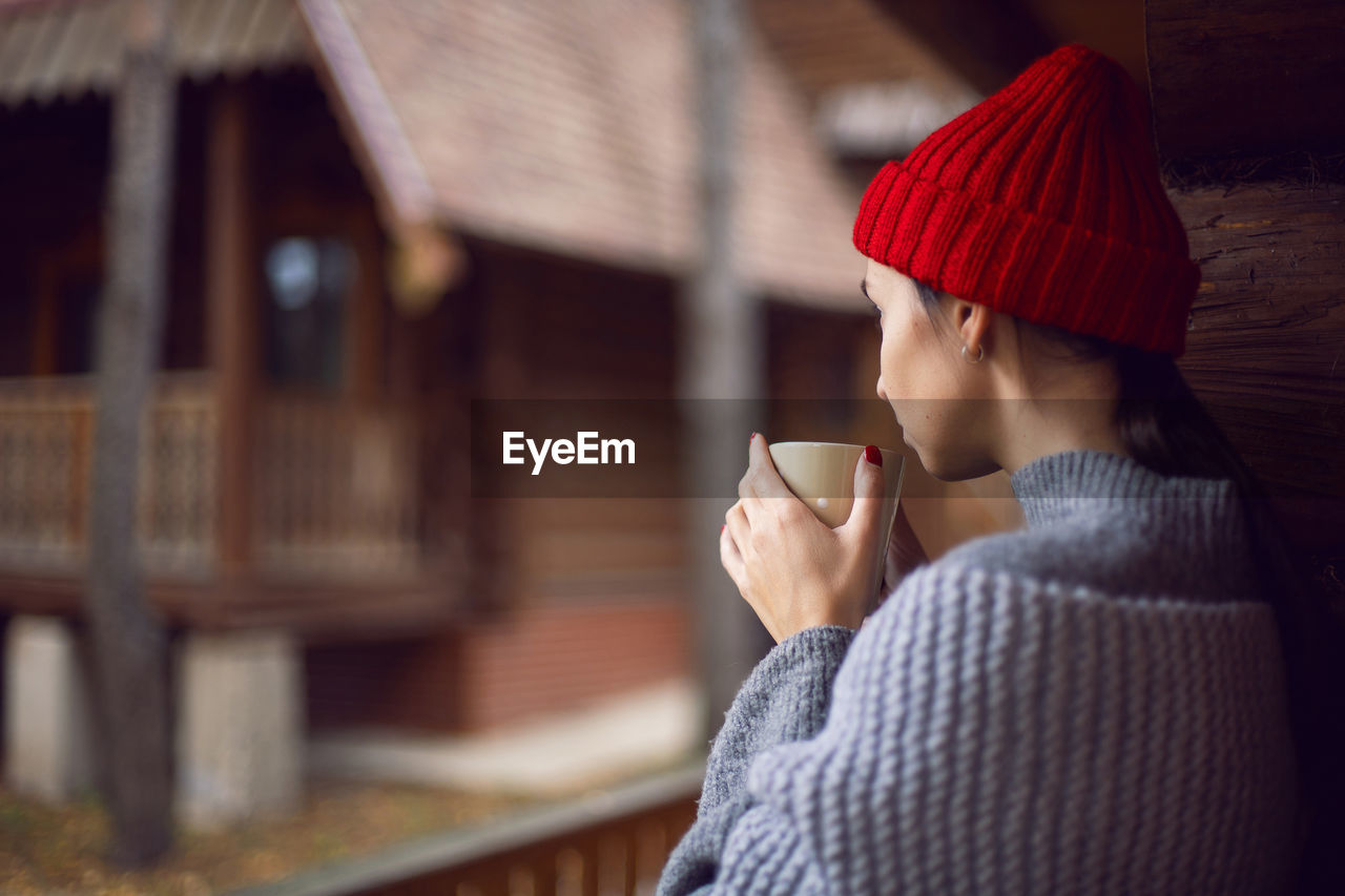 Woman in a red hat and scarf and a mug stands at a wooden house, in the woods in the autumn morning 