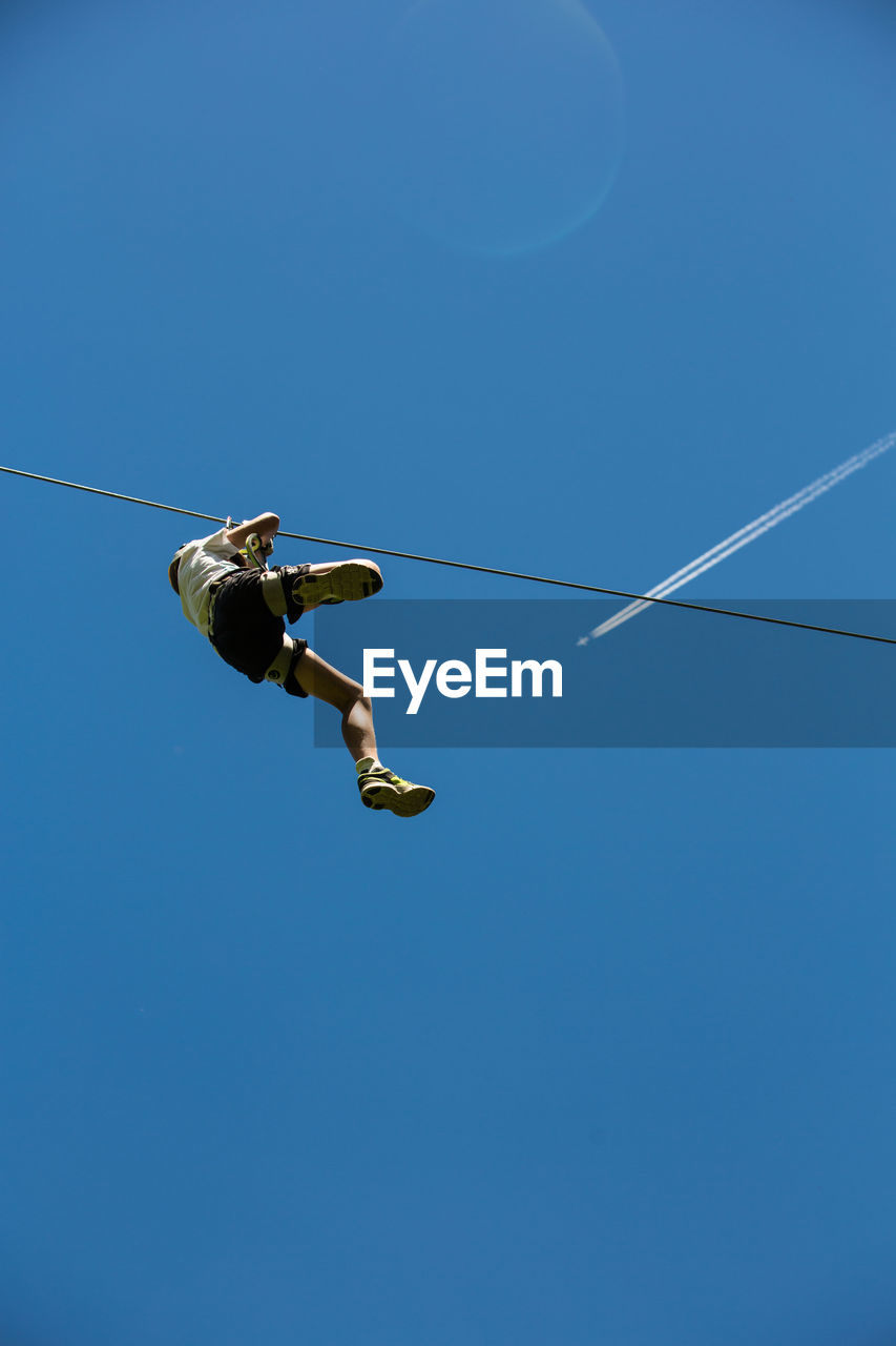 Low angle view of teenage boy zip lining against clear blue sky during sunny day