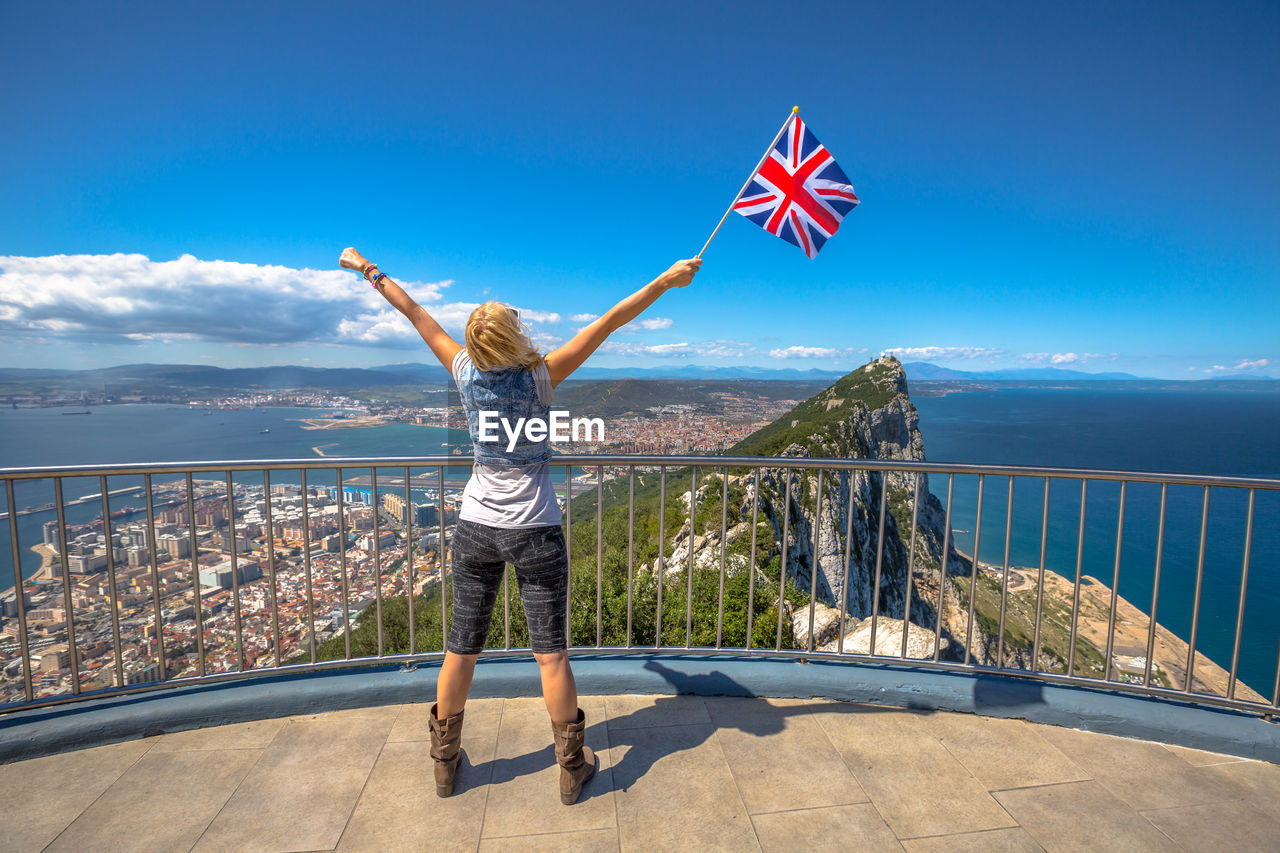 Woman holding flag while standing by railing against sky