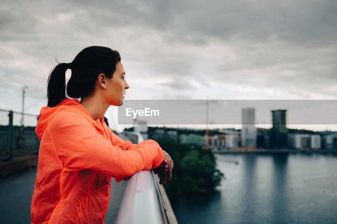 Side view of female athlete in raincoat looking away while standing on footbridge over sea