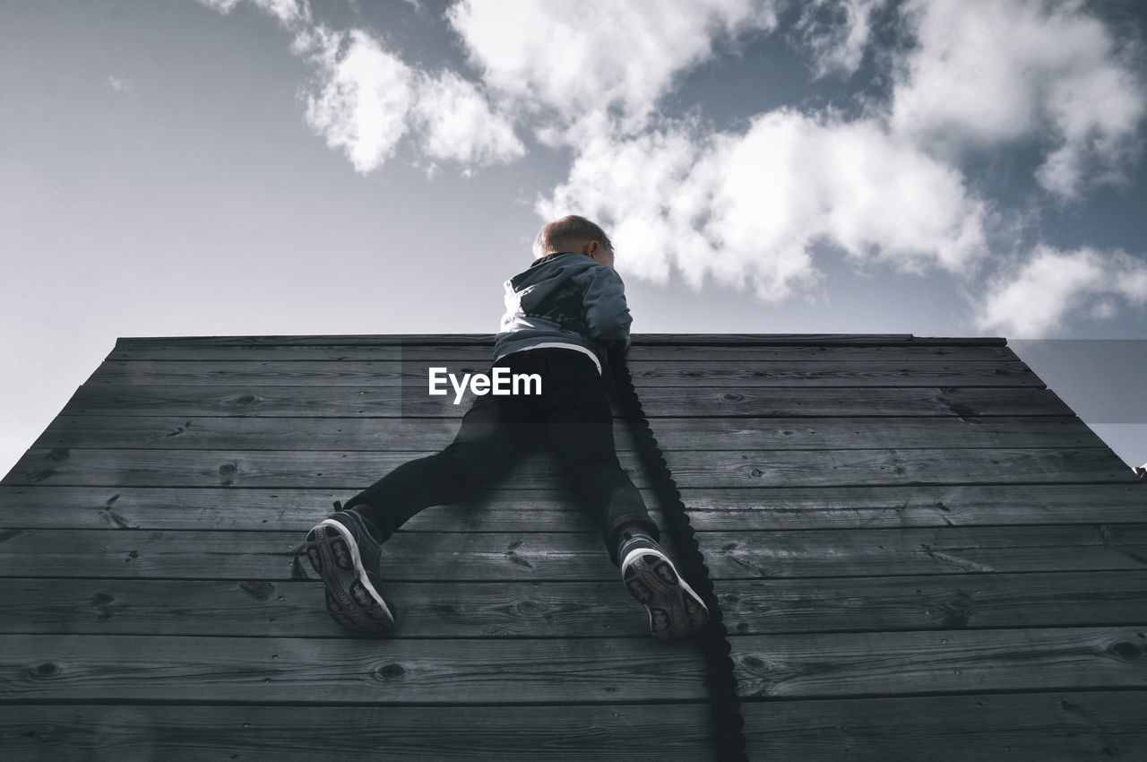 Low angle view of boy sitting on wood against sky