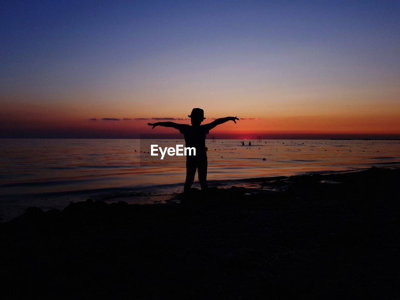 Silhouette boy standing with arms outstretched at beach against sky during sunset