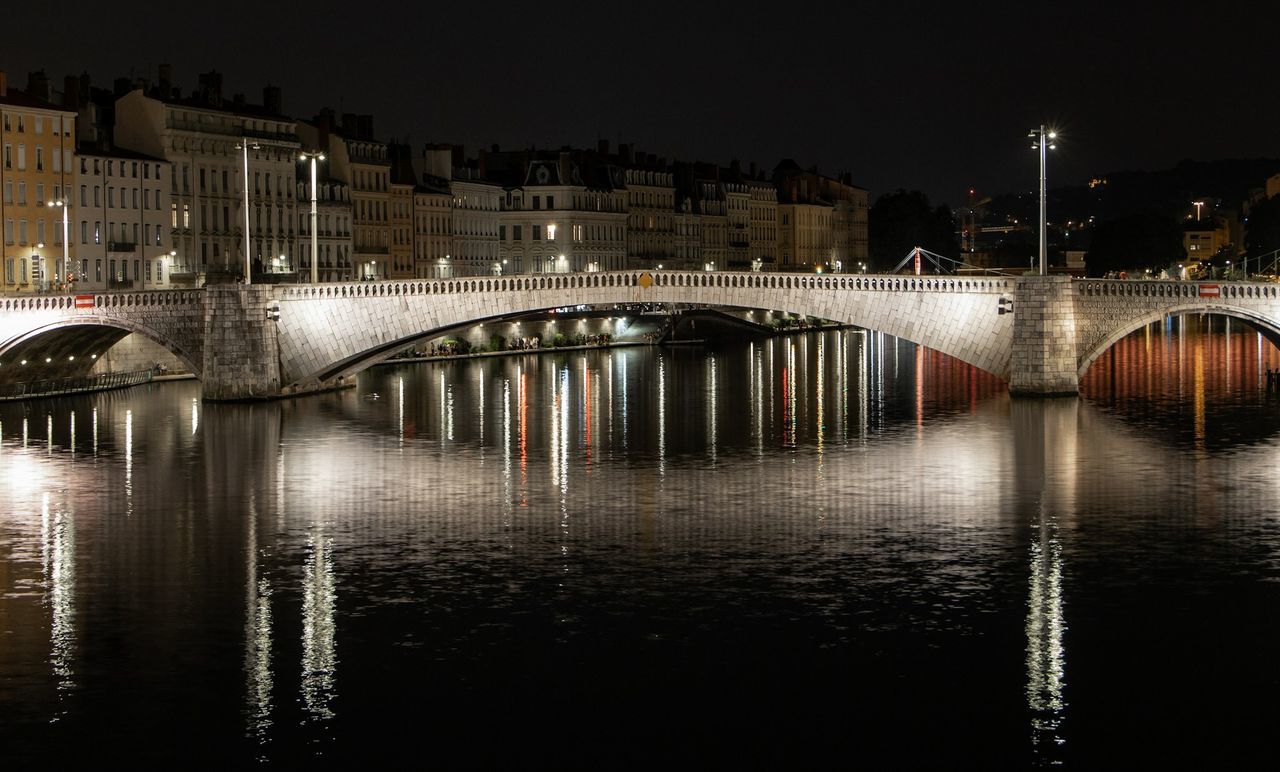 BRIDGE OVER RIVER BY ILLUMINATED BUILDINGS AT NIGHT