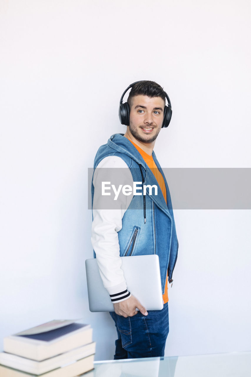 Young man looking away while holding laptop against white wall