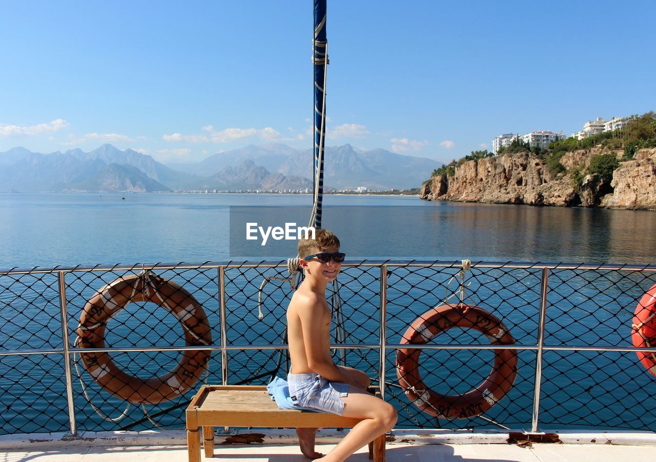 Shirtless boy sitting in boat by river against sky during sunny day