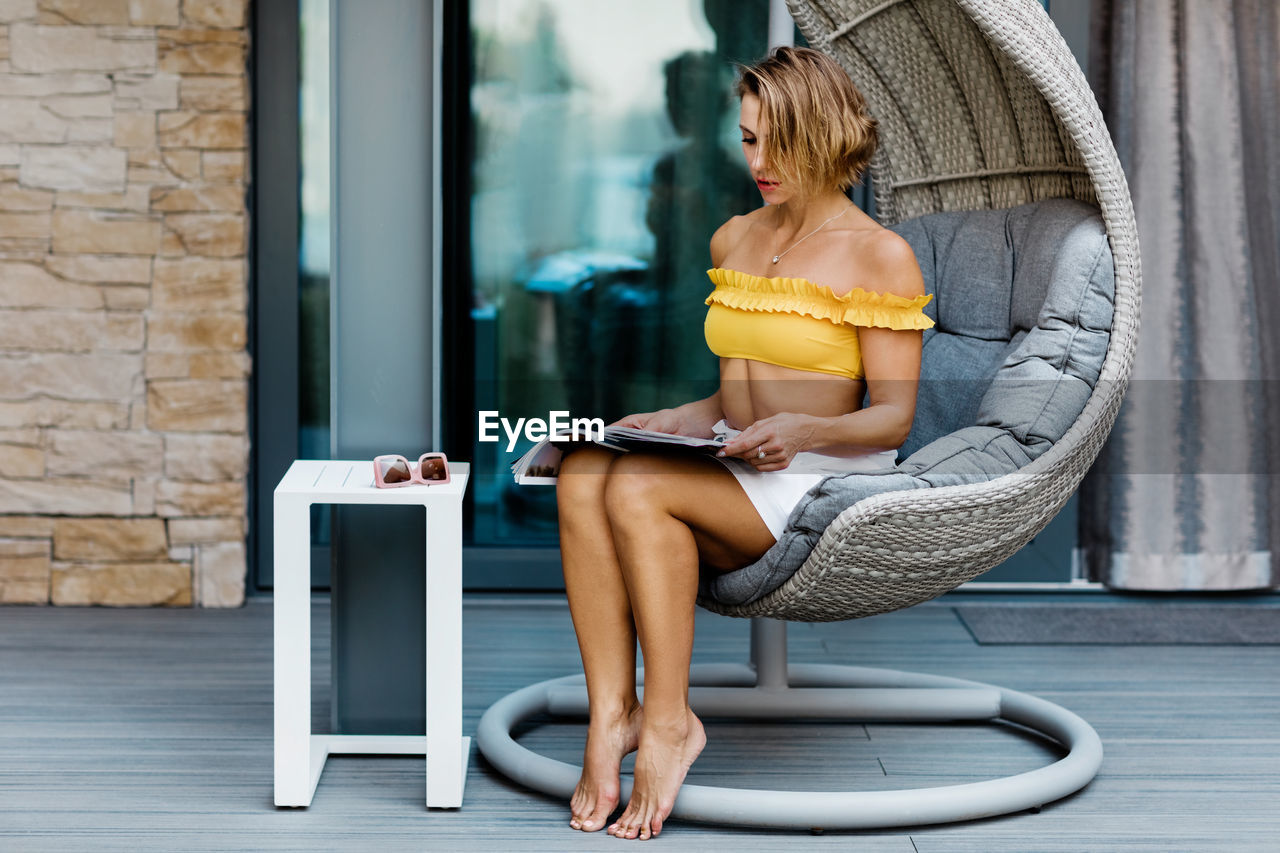 A young woman is sitting in a hanging chair on the terrace and reading a magazine