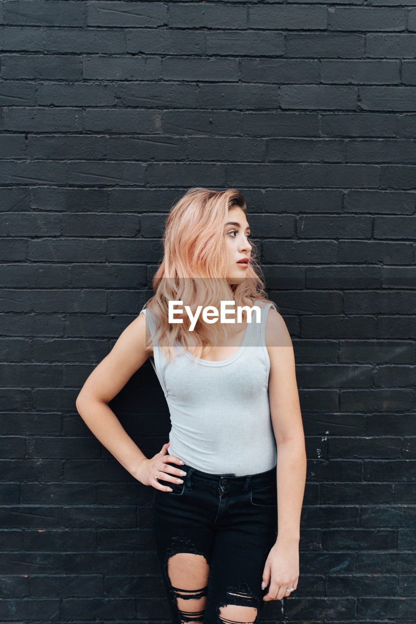 Young woman looking away while standing with hand on hip against black brick wall