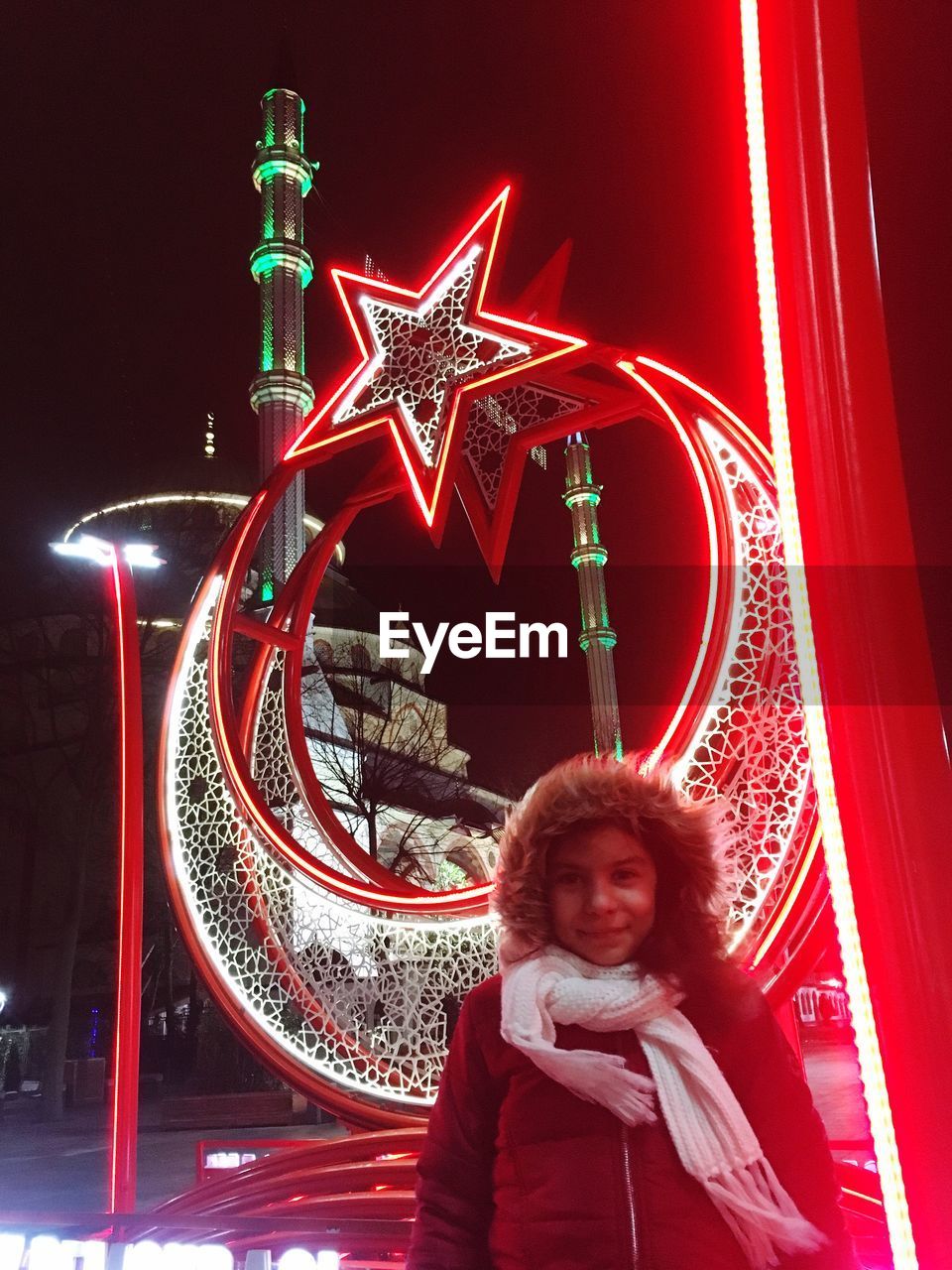 PORTRAIT OF SMILING YOUNG WOMAN STANDING IN ILLUMINATED AMUSEMENT PARK