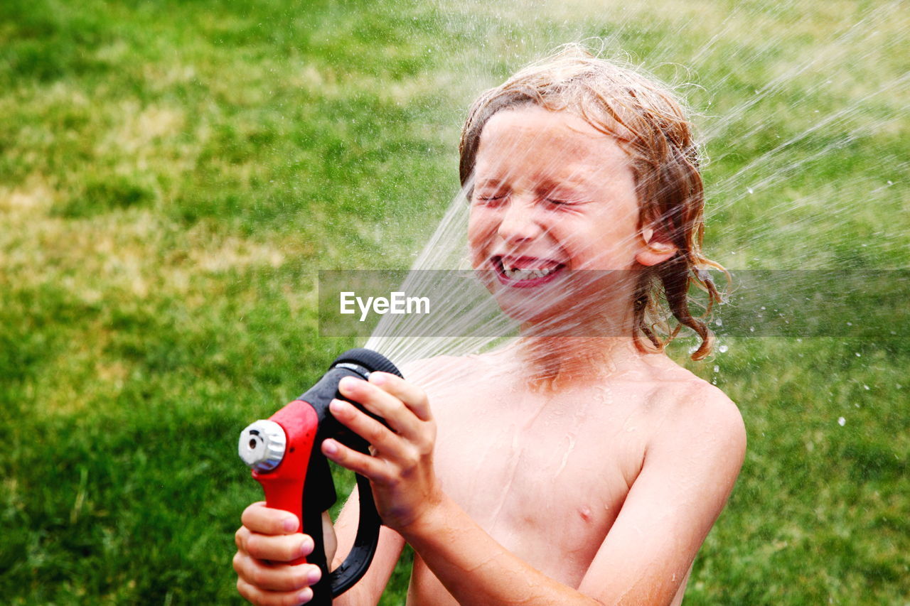 Shirtless boy splashing water from garden hose on face in back yard