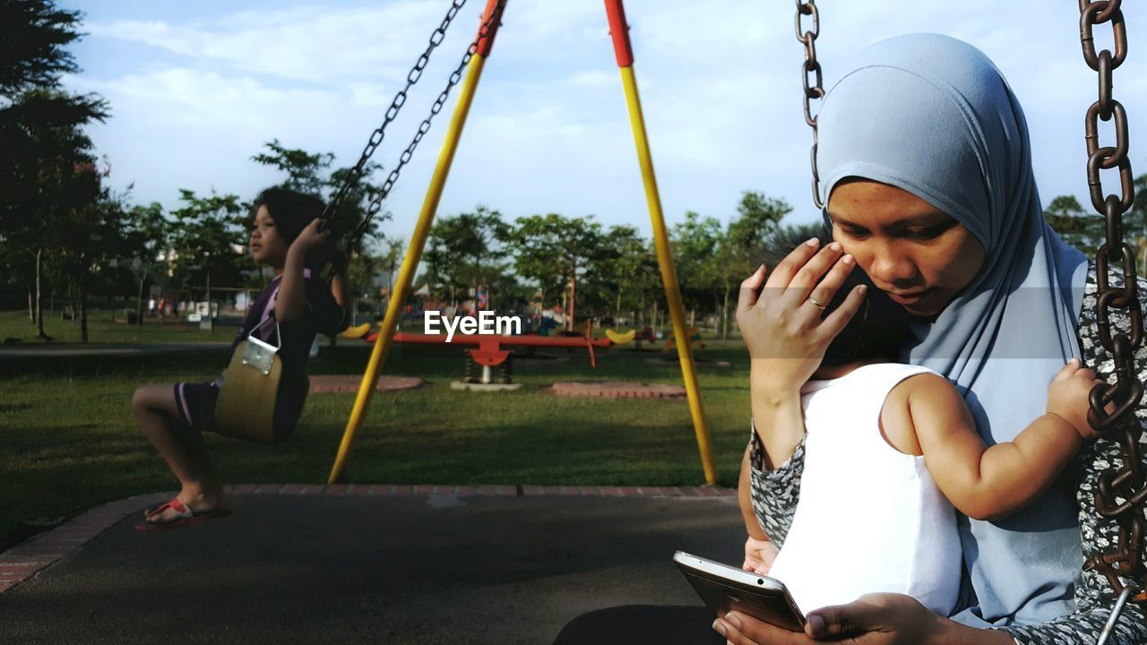 Woman sitting on swing with daughter while using mobile phone at park