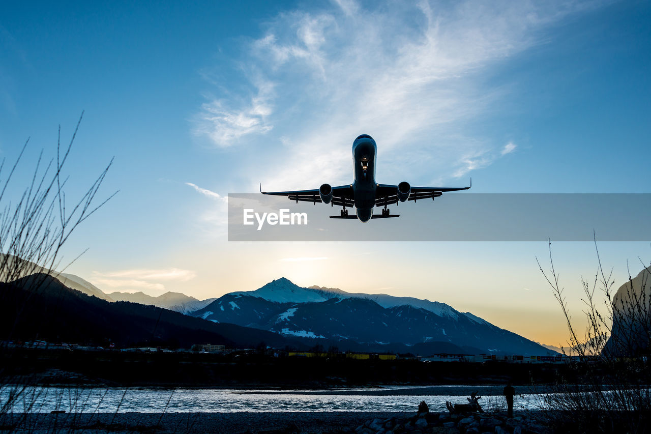 AIRPLANE FLYING IN SKY OVER SNOWCAPPED MOUNTAINS AGAINST CLOUDY BLUE