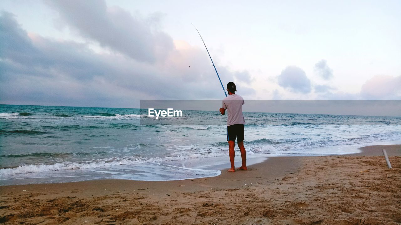 Rear view of young man fishing on beach against sky