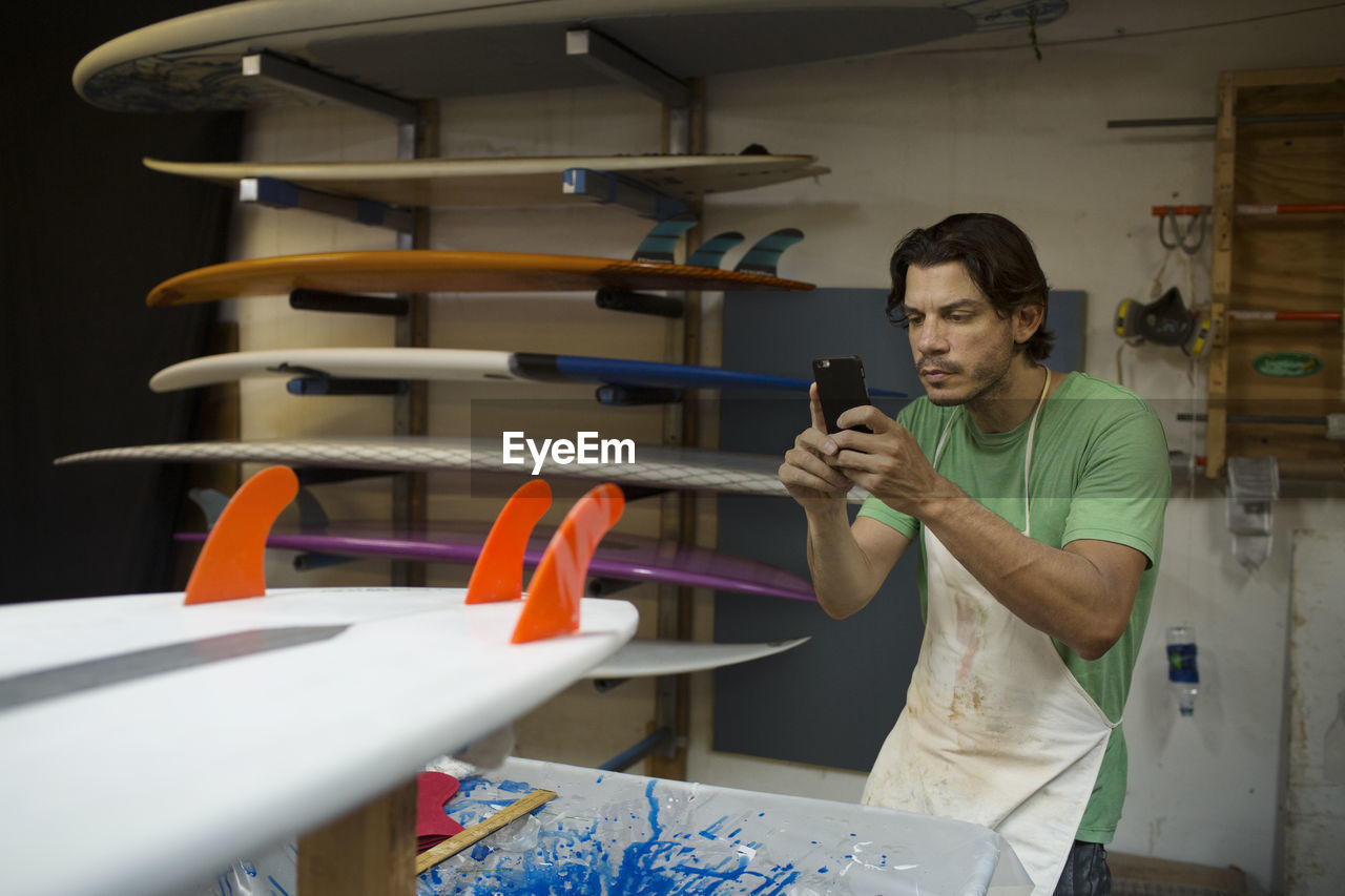 Male worker photographing surfboard through smart phone in workshop