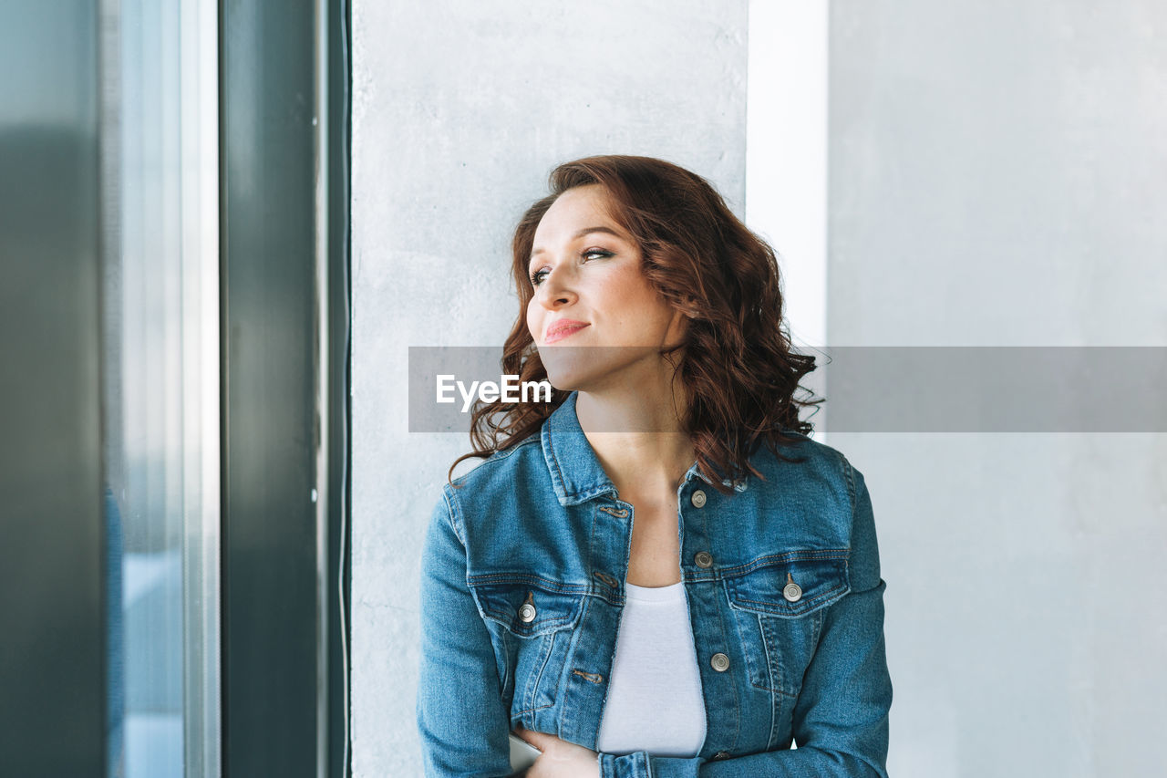 Portrait of smiling charming young woman in jeans jacket near window in modern building