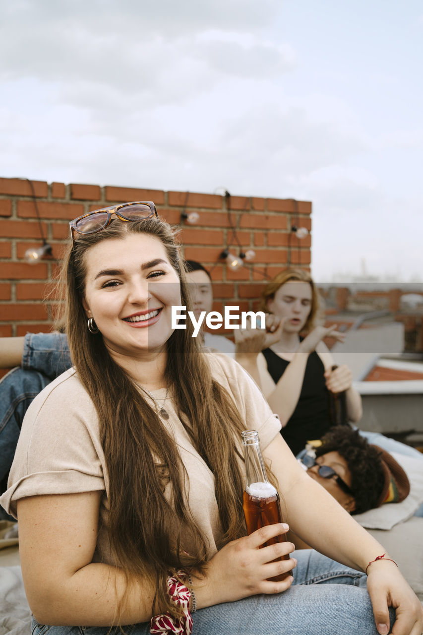 Portrait of smiling young woman holding beer bottle sitting in front of friends on rooftop