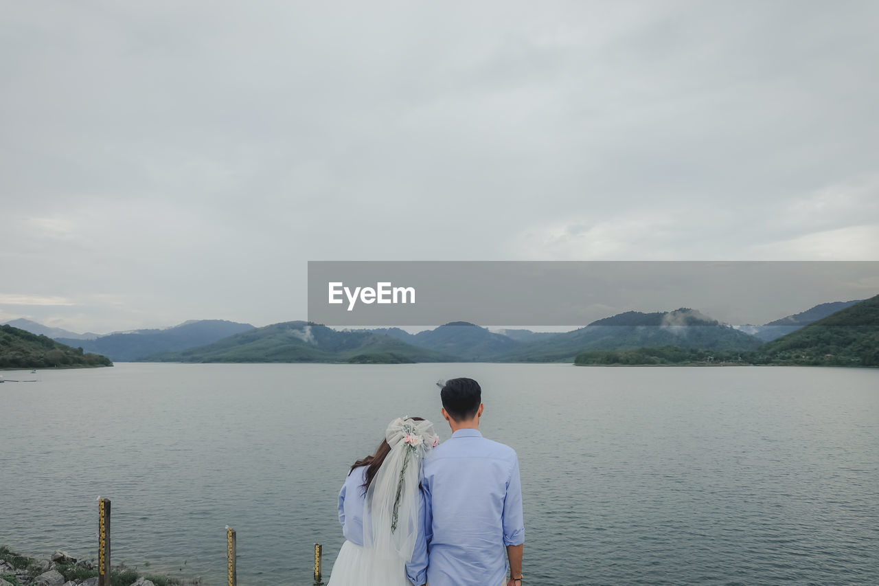 REAR VIEW OF MAN LOOKING AT LAKE AGAINST MOUNTAIN