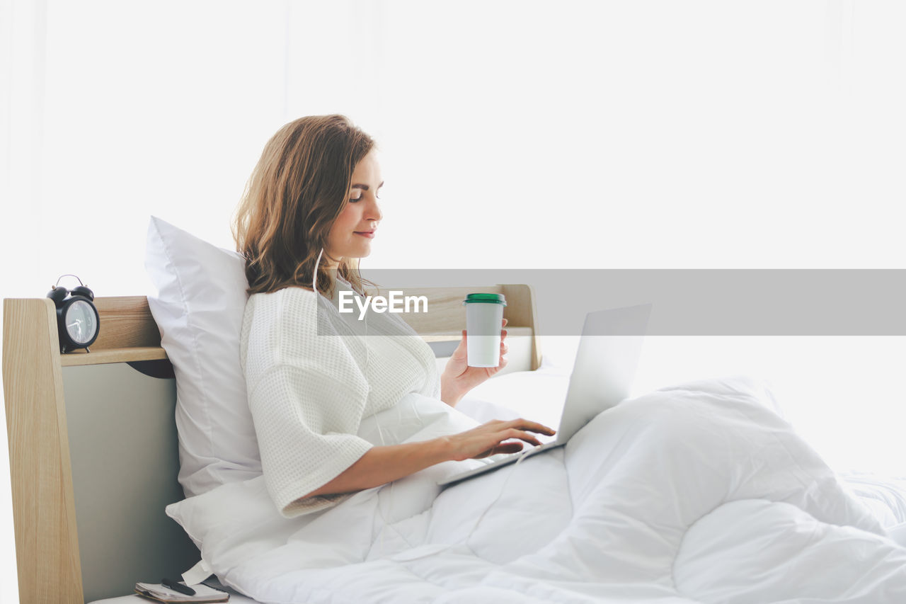 Woman using laptop while holding coffee cup on bed