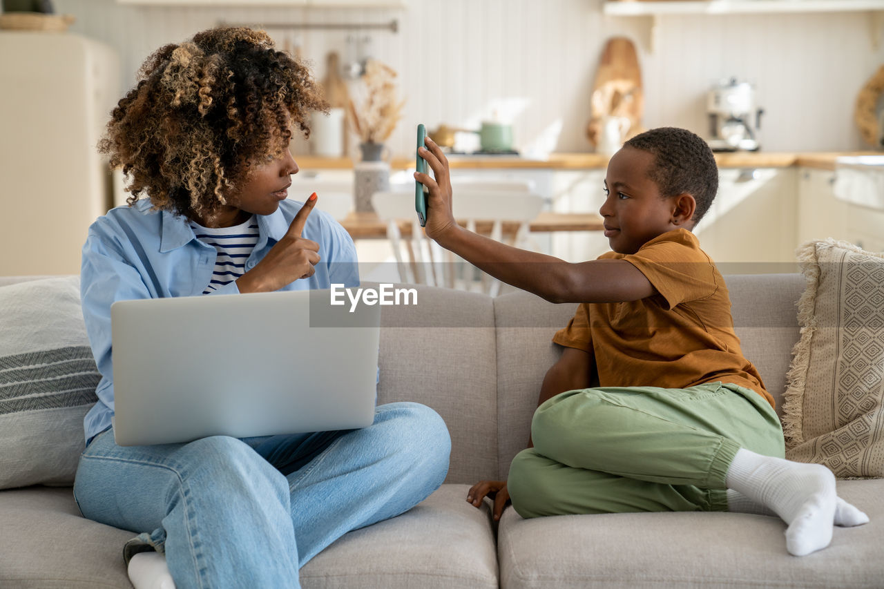 Freelancer black mother working on laptop at home, noisy african child distracts asking attention.