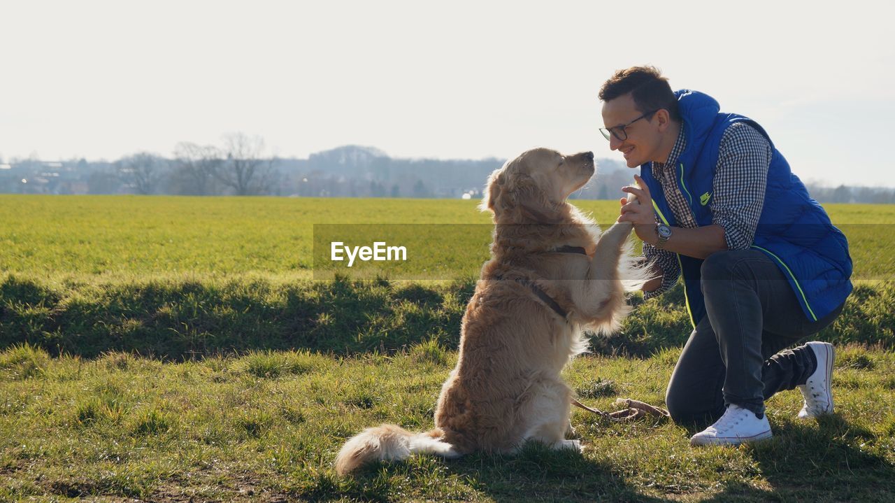 Mid adult man playing with dog while kneeling on grassy field against sky