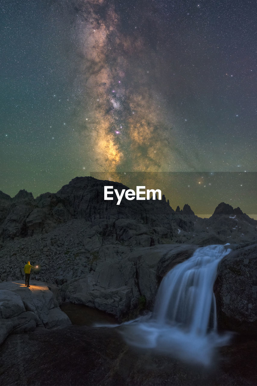 Anonymous traveler with torch contemplating waterfall streaming among rough rocky terrain under night starry sky with bright glowing milky way person