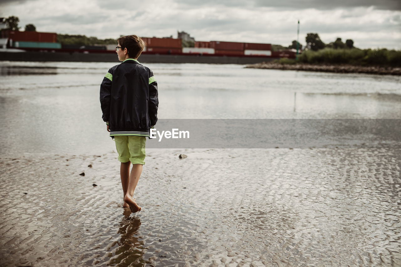 Young boy walk on vacation at mud flat shore and watch a passing container ship