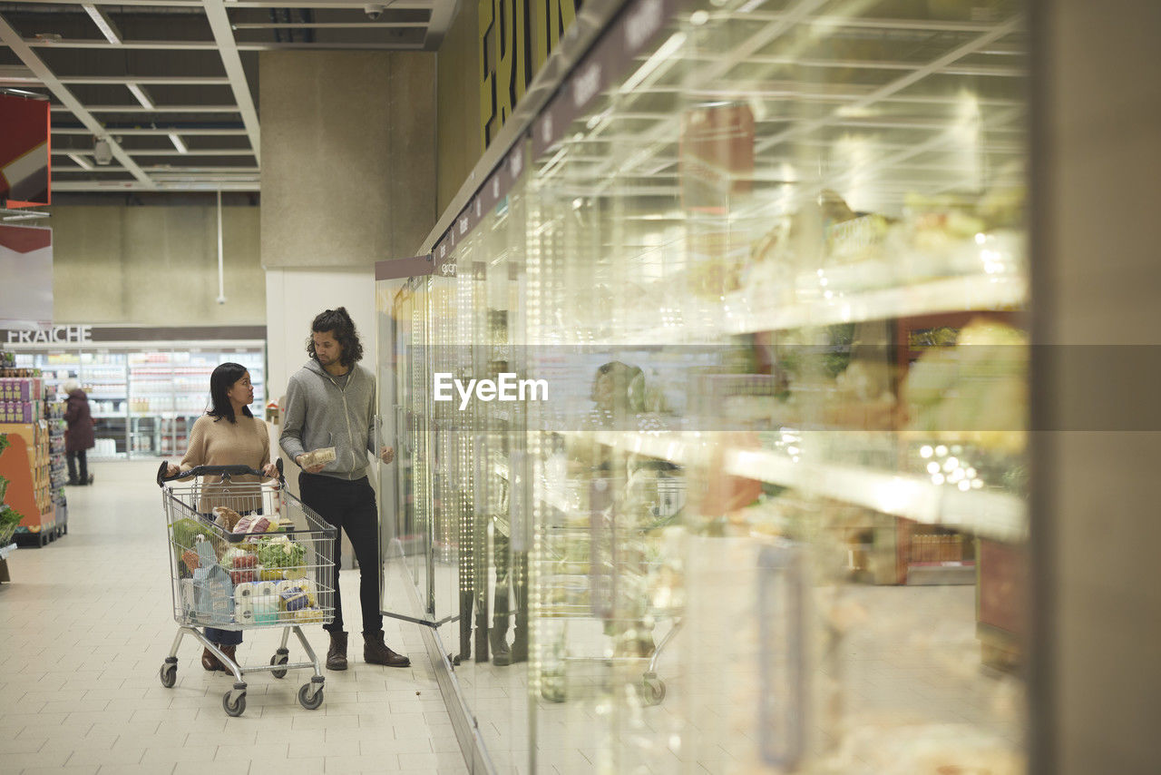 Couple in supermarket talking in front of fridges with vegetables