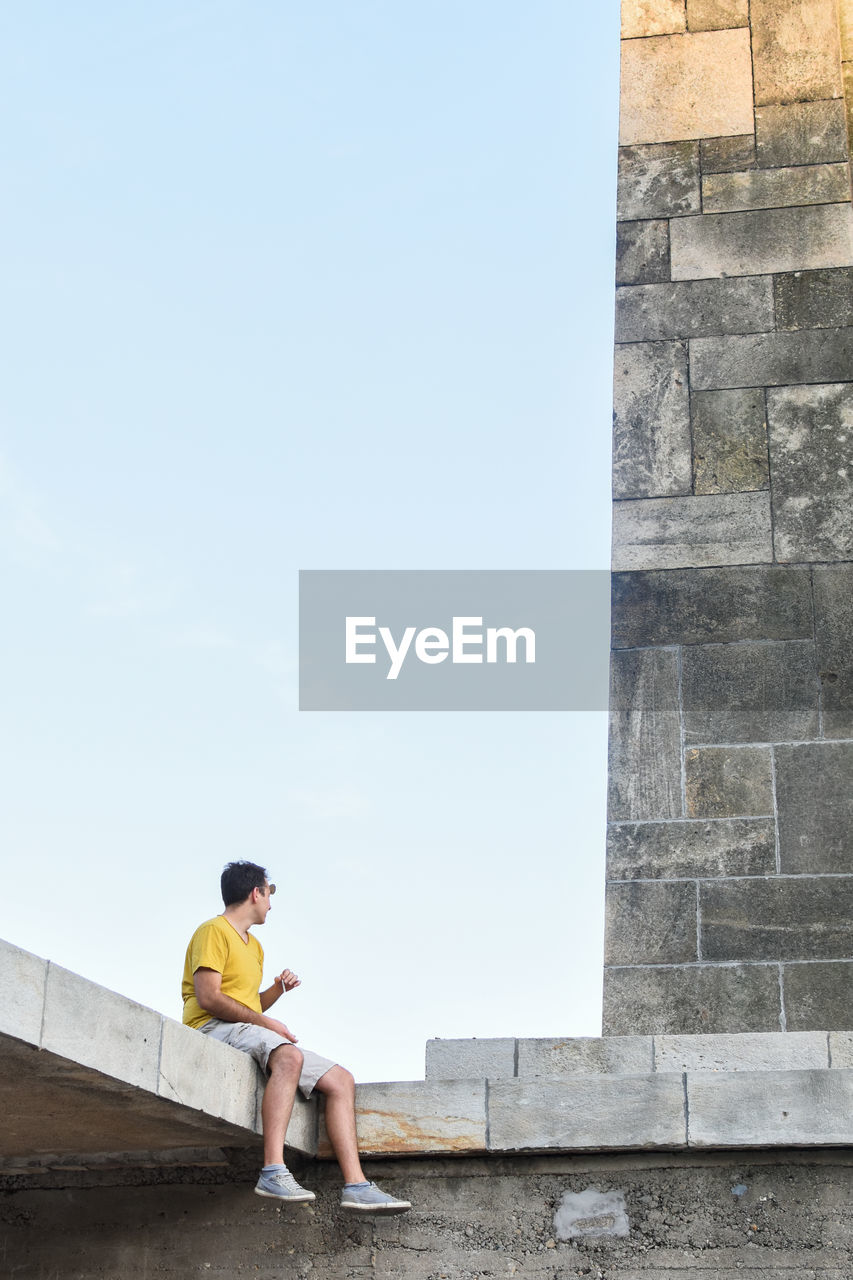 REAR VIEW OF BOY SITTING ON STONE WALL