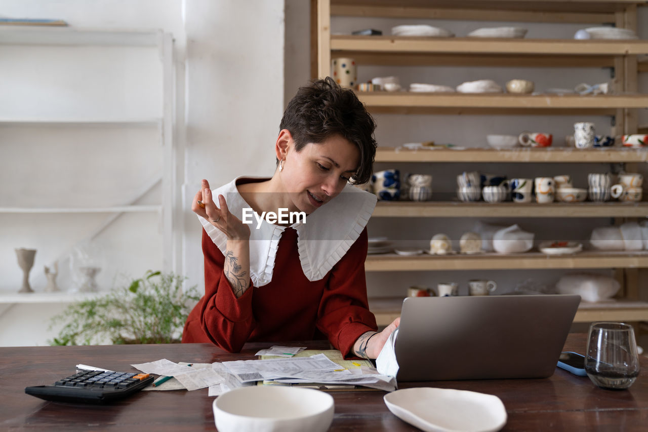 Focused young woman ceramic studio owner analyzing bills and receipts, doing pottery cost analysis