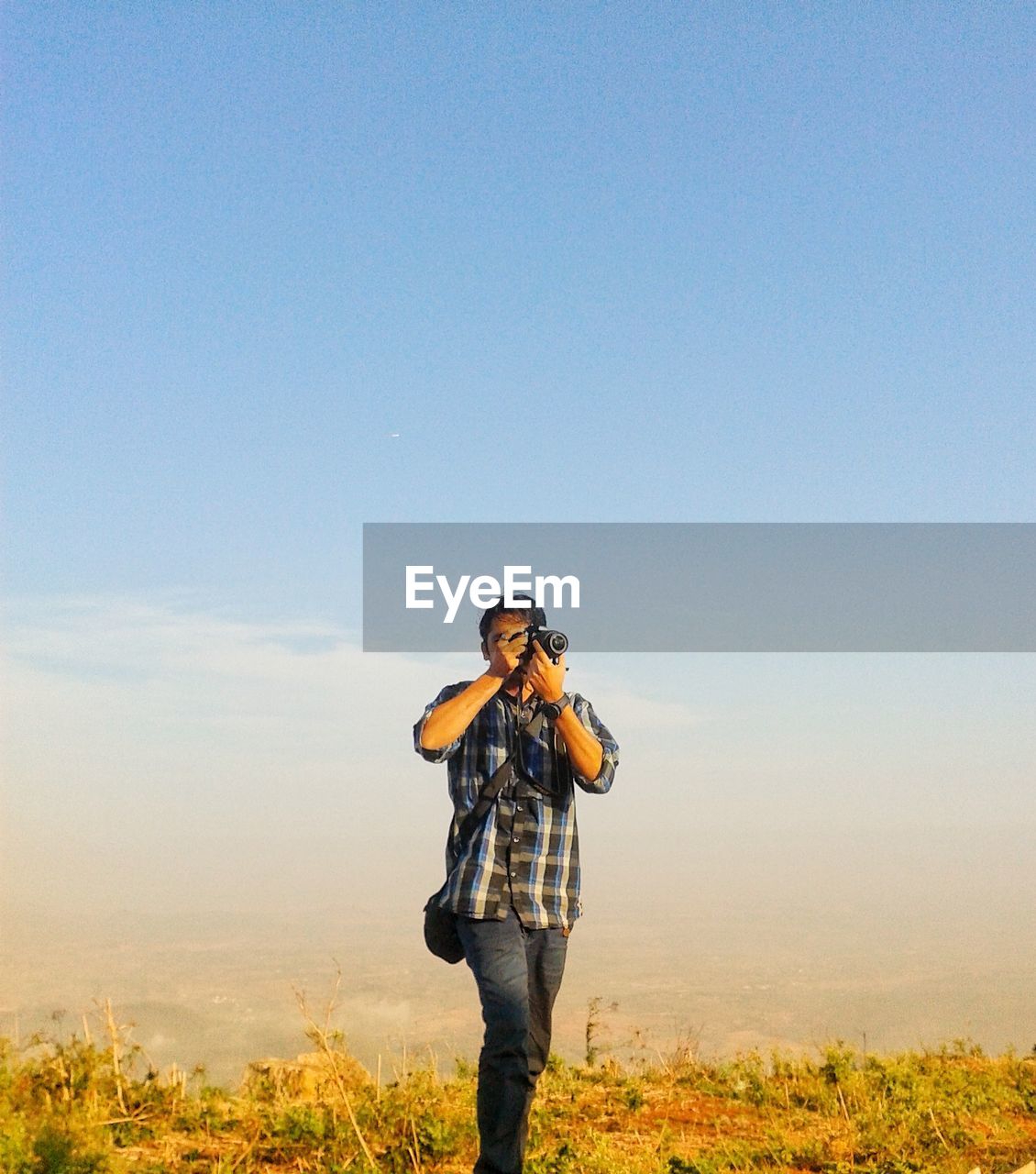 Young man photographing with digital camera on field against sky during sunset