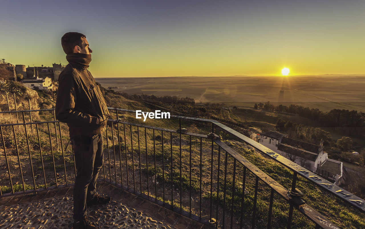 Side view of man looking at landscape while standing by railing during sunset