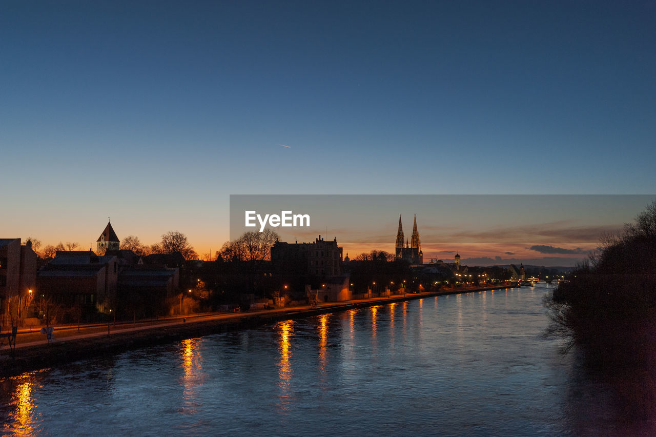 RIVER BY ILLUMINATED BUILDINGS AGAINST CLEAR SKY AT SUNSET