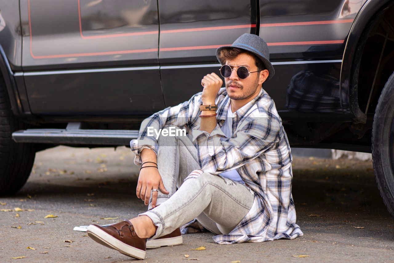 Full length portrait of man wearing goggles sitting on road by car