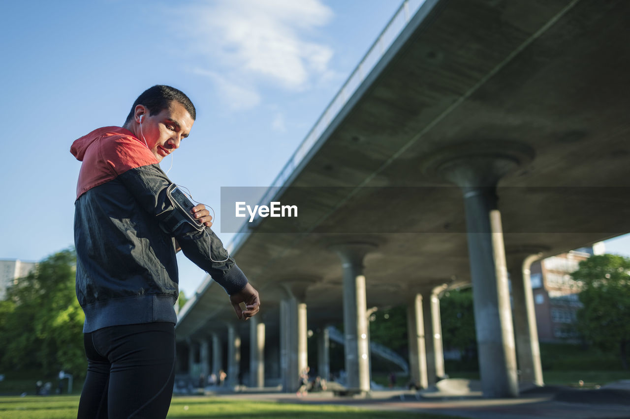 Low angle view of man touching arm band while standing against bridge