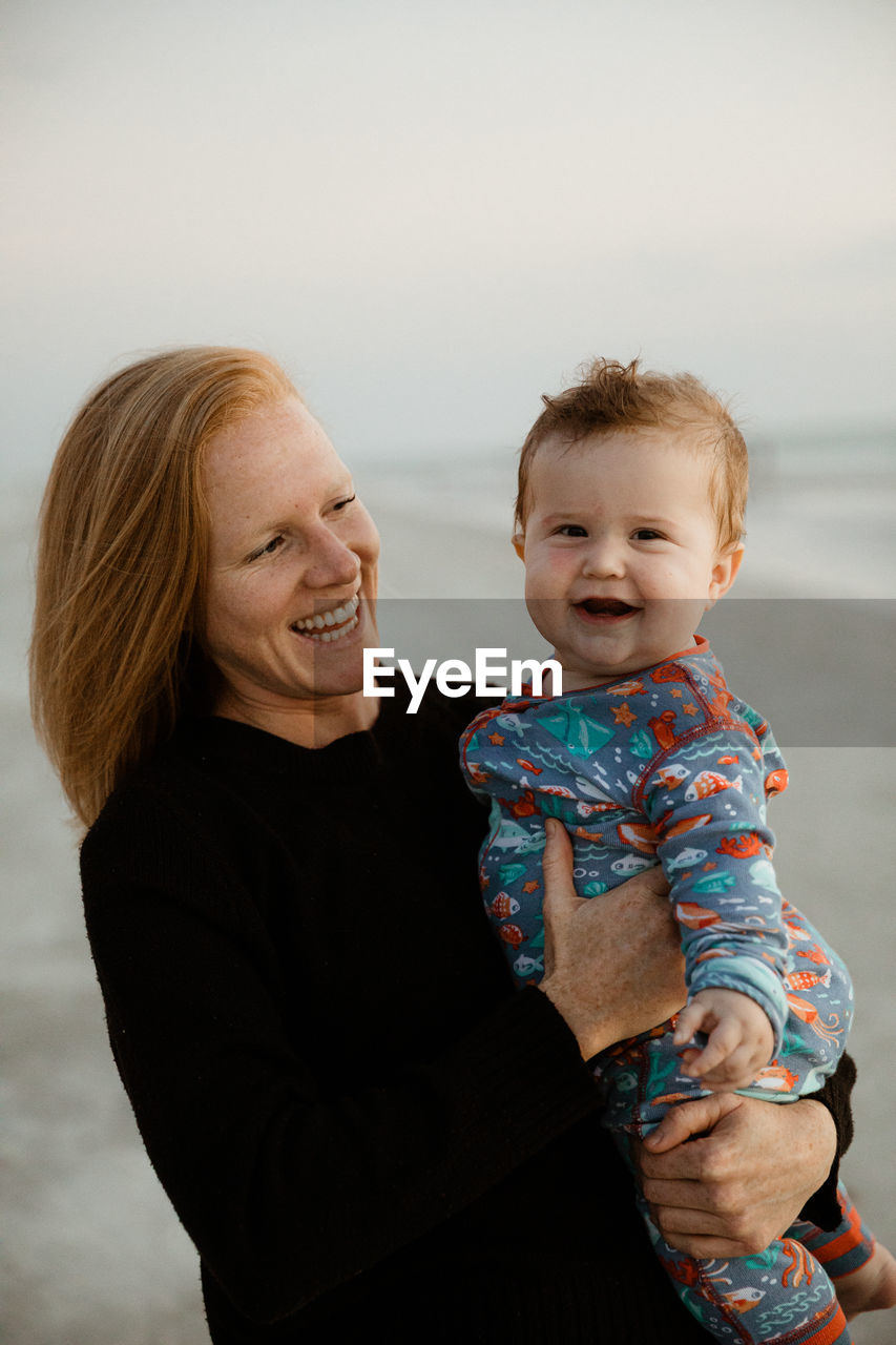 Young redhead mom laughs and holds laughing fat baby boy in onesie
