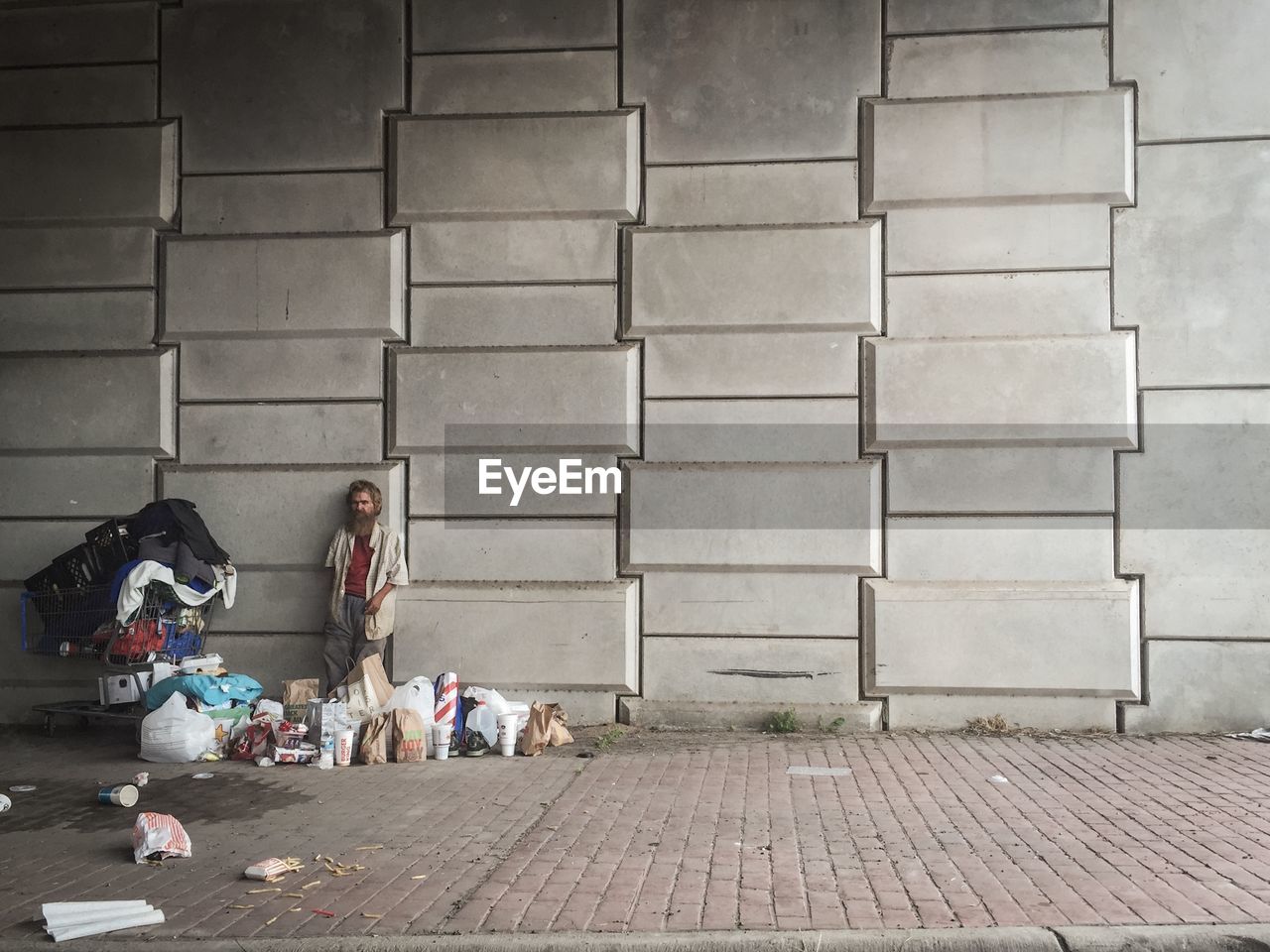 Homeless man standing by shopping cart and bags against wall